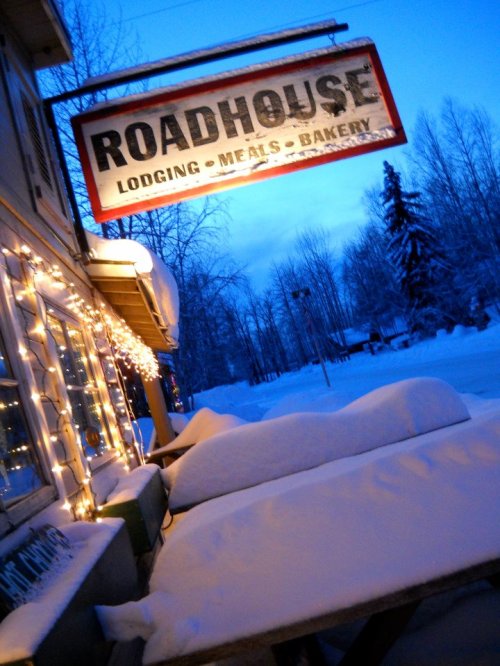 A cozy roadhouse sign illuminated at dusk, surrounded by snow-covered tables and trees.