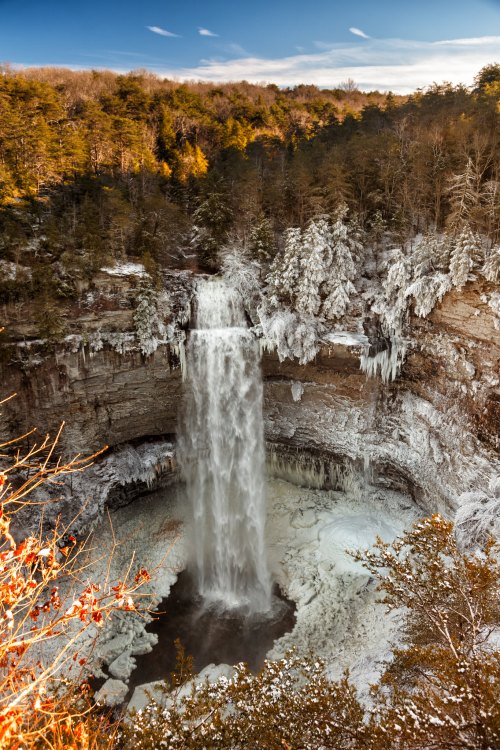 A stunning waterfall cascades into a frozen pool, surrounded by snow-covered trees and a winter landscape.