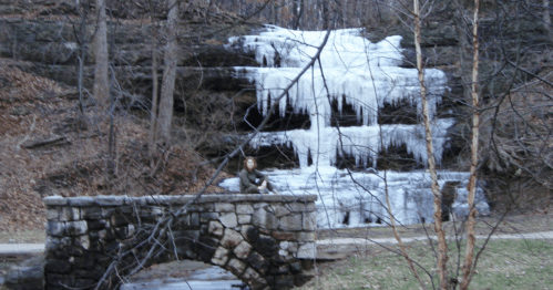 A stone bridge in a forest with a frozen waterfall and a person sitting on the bridge, surrounded by bare trees.