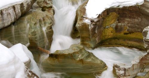 A serene waterfall cascades over smooth, layered rocks, surrounded by patches of snow.