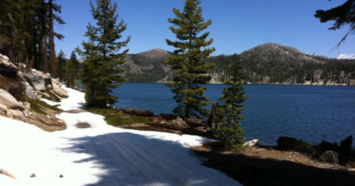 A serene lake surrounded by mountains and pine trees, with patches of snow along a walking path.
