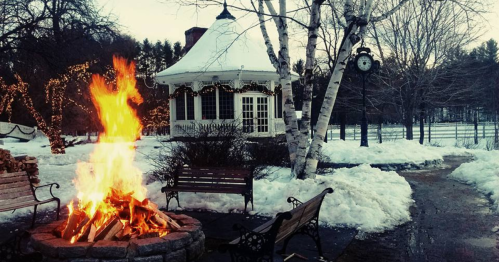 A cozy winter scene featuring a fire pit, snow-covered ground, a gazebo, and a clock surrounded by bare trees.