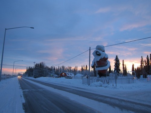 A snowy road with a large Santa statue and trees under a colorful sunset sky.