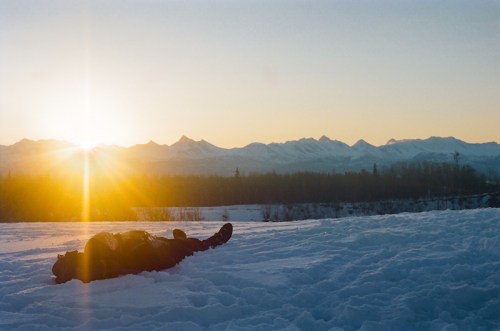 A person lying in the snow, with a sunset illuminating the snowy mountains in the background.