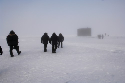 A group of people in winter gear walking across a snowy landscape in foggy conditions, with a structure in the background.