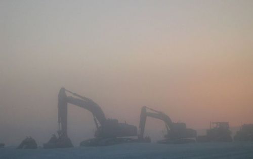 Silhouettes of excavators in a foggy landscape at dawn, with soft pastel colors in the sky.