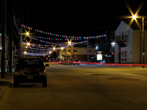 A quiet street at night adorned with colorful string lights, with a parked car and light trails from passing vehicles.