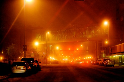 A foggy street scene illuminated by orange streetlights, with a pedestrian bridge in the background and parked cars on the side.
