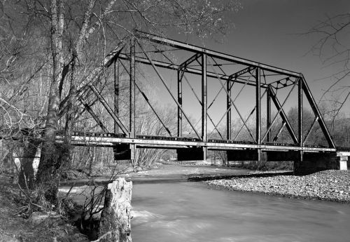 Black and white image of a metal truss bridge over a flowing river, surrounded by bare trees and rocky banks.