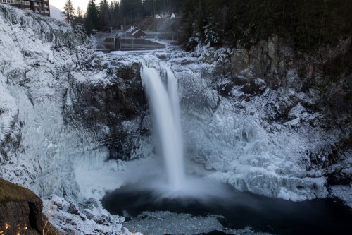A majestic waterfall cascades over icy rocks, surrounded by snow-covered trees and a serene winter landscape.