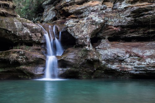 A serene waterfall cascades over rocky cliffs into a clear, turquoise pool surrounded by lush greenery.