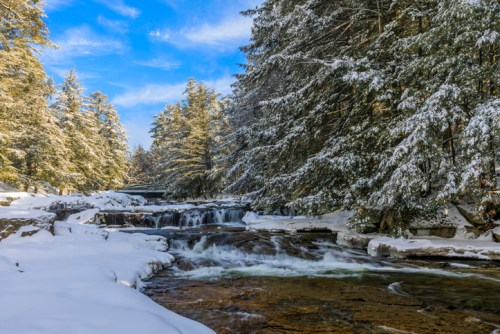 A serene winter landscape featuring a flowing river surrounded by snow-covered trees and a clear blue sky.