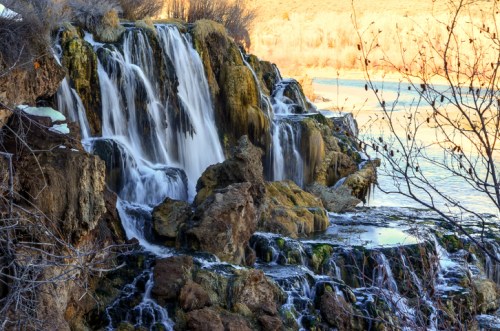 A serene waterfall cascading over rocks, surrounded by greenery and reflecting sunlight on a calm river.