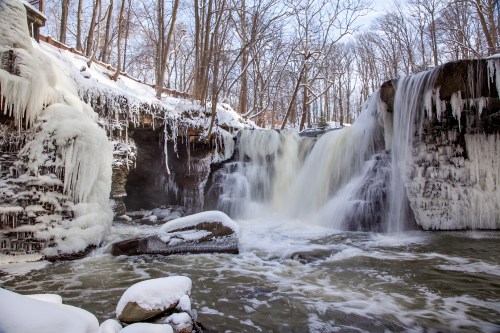 A frozen waterfall surrounded by snow-covered rocks and trees, with icicles hanging from the cliff edges.