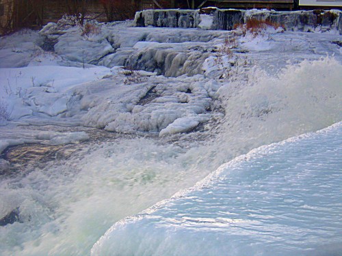 A frozen river with icy banks and splashing water, surrounded by snow-covered rocks and winter scenery.