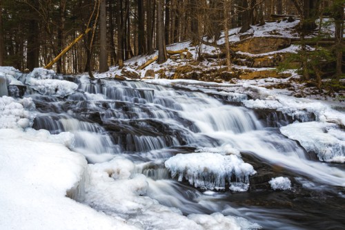A serene waterfall cascades over icy rocks, surrounded by trees in a winter landscape.
