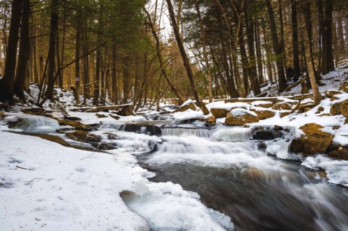 A serene winter scene of a flowing stream surrounded by snow-covered rocks and trees in a forest.