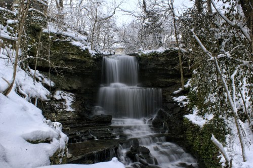 A serene waterfall cascading over rocks, surrounded by snow-covered trees and a winter landscape.