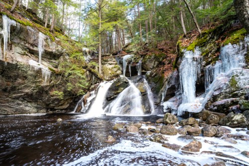 A serene waterfall cascades over rocks, surrounded by lush greenery and icicles, reflecting a tranquil natural scene.