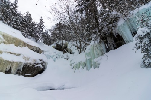 A snowy landscape featuring icy cliffs and frozen waterfalls, surrounded by trees under a cloudy sky.