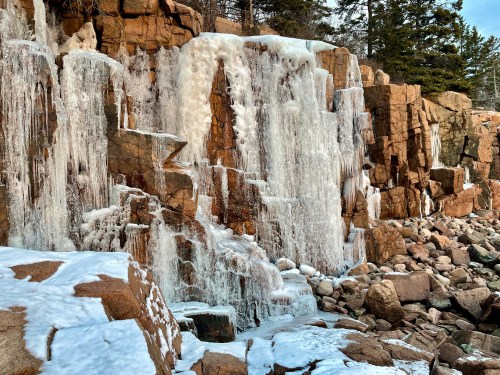 Frozen waterfall cascading down rocky cliffs, surrounded by snow and boulders, with evergreen trees in the background.