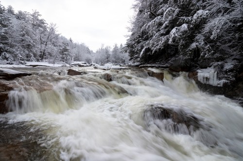 A snowy landscape featuring a rushing river with frosty rocks and trees, creating a serene winter scene.