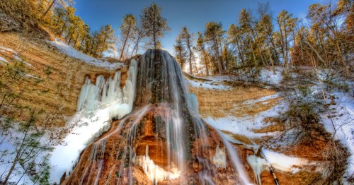 A frozen waterfall cascading down a rocky cliff, surrounded by snow and tall trees under a clear blue sky.