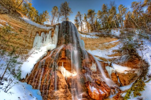 A stunning waterfall cascading down a rocky cliff, surrounded by snow and evergreen trees under a clear blue sky.