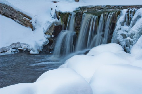 A serene winter scene featuring a waterfall cascading over rocks, surrounded by snow-covered terrain.