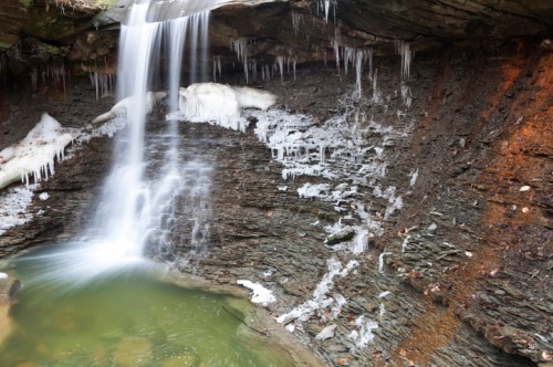 A serene waterfall cascades over rocky cliffs, surrounded by icicles and a calm pool of water below.