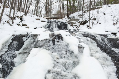 A snowy waterfall cascades over dark rocks, surrounded by bare trees and a wooden bridge in the background.