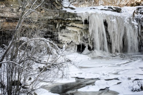 A frozen waterfall surrounded by snow-covered rocks and trees, with icicles hanging from the cliff above.