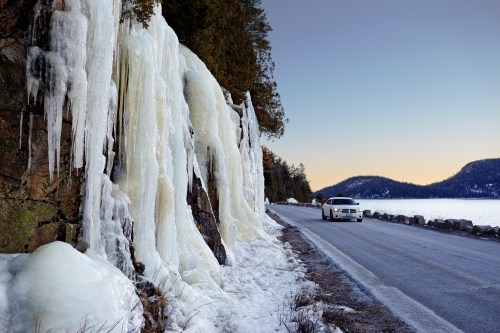 A car drives along a road beside icy rock formations and a frozen lake at sunset.