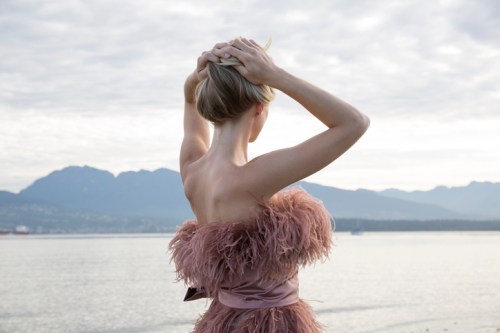 A woman in a feathered dress stands by the water, facing away, with mountains in the background and a cloudy sky.
