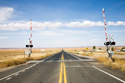 A wide, empty road stretches into the distance, flanked by railroad crossing gates against a clear blue sky.