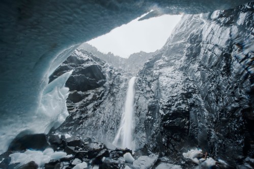 A waterfall cascades down rocky cliffs, viewed from an icy cave entrance, with mist and dark stone surrounding the scene.
