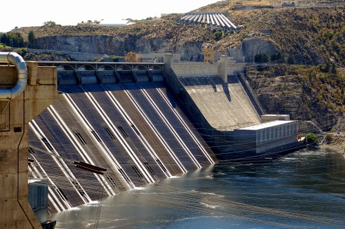 A large dam with a sloped concrete face, surrounded by rocky terrain and water, reflecting sunlight.
