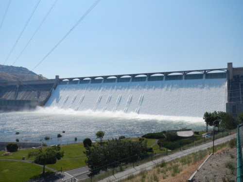 A large dam releasing water, creating a cascade over the structure, with greenery and pathways in the foreground.