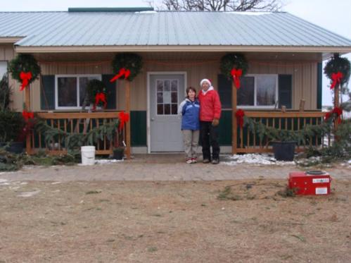 Two people stand in front of a decorated house with wreaths and red bows, surrounded by a snowy landscape.