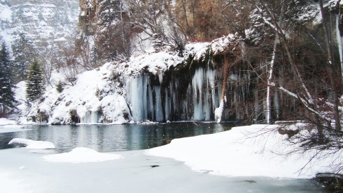 A snowy landscape featuring a frozen waterfall beside a calm, icy lake surrounded by trees and rocky cliffs.