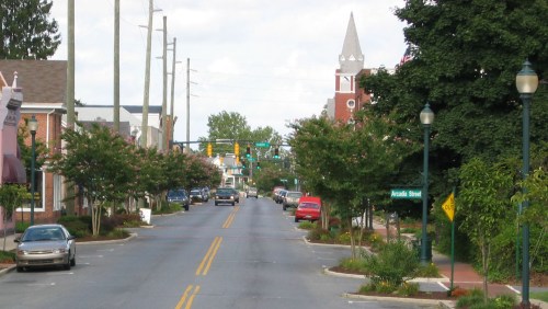 A quiet street lined with trees, featuring traffic lights, parked cars, and a church steeple in the background.