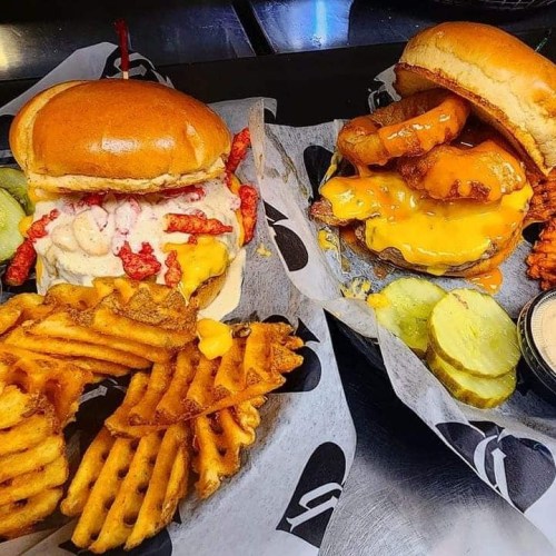 Two gourmet burgers with toppings, served with waffle fries and pickles on the side, all on a black and white paper.