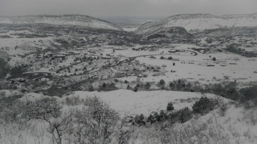 A snowy landscape with rolling hills, trees, and a winding road, under a cloudy sky.