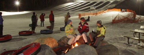 A group of people sitting around a fire pit at a snow tubing park, with illuminated slopes in the background.