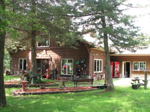 A rustic wooden house surrounded by trees, featuring large windows and a porch with benches and garden decor.