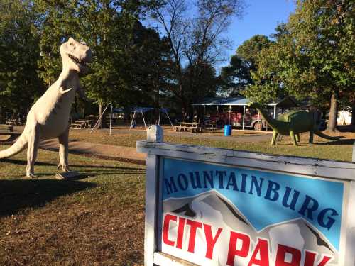 A dinosaur statue stands near a sign for Mountainburg City Park, with playground equipment and trees in the background.