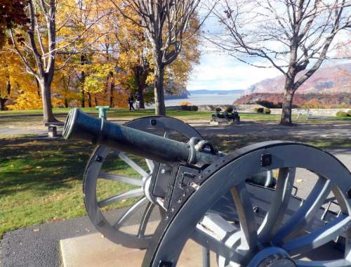 A historic cannon in a park, surrounded by colorful autumn trees and a scenic view of a lake and mountains in the background.