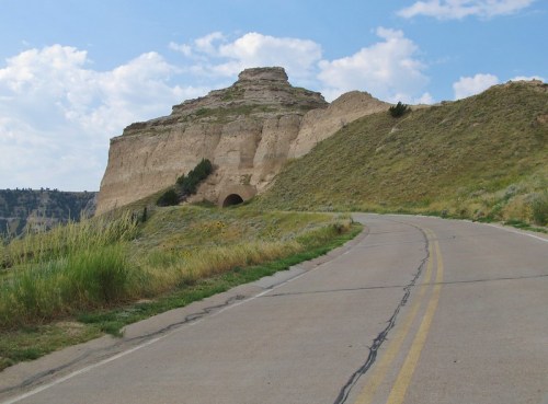 A winding road leads towards a rocky hill with a tunnel entrance, under a blue sky with scattered clouds.