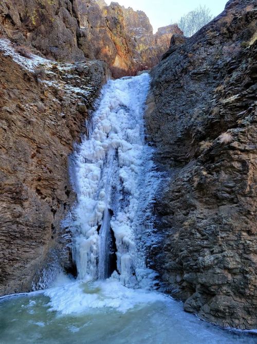 A frozen waterfall cascading down rocky cliffs, surrounded by ice and snow, with a small pool at the base.