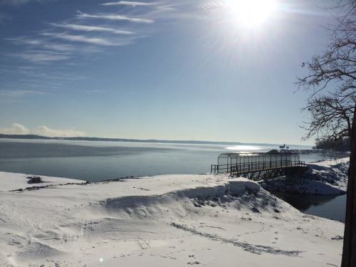 A serene winter landscape featuring a snowy shore, calm water, and a distant dock under a bright sun.